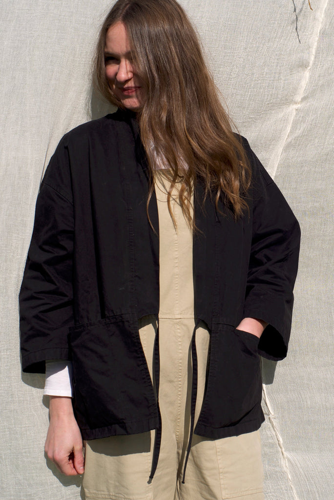 A female wearing a heavy organic cotton drill garden kimono, in dexter black whilst posing with one of her hands in the bar tacked pocket