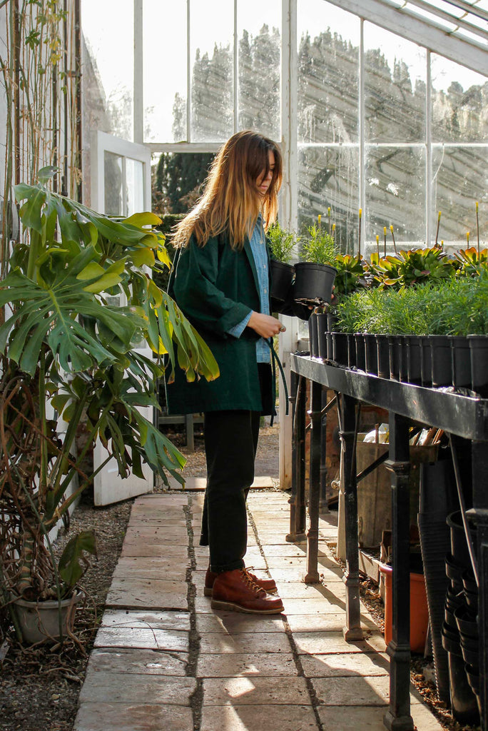 A female wearing a heavy organic cotton drill garden kimono, in laurel green whilst holding a plant in a greenhouse